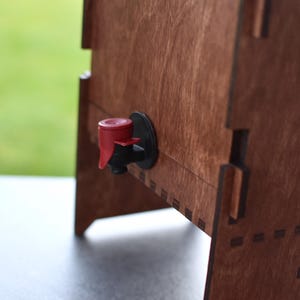 Puede incluir: Un dispensador de madera con un grifo rojo y negro. El dispensador est&aacute; hecho de madera y tiene un acabado natural.