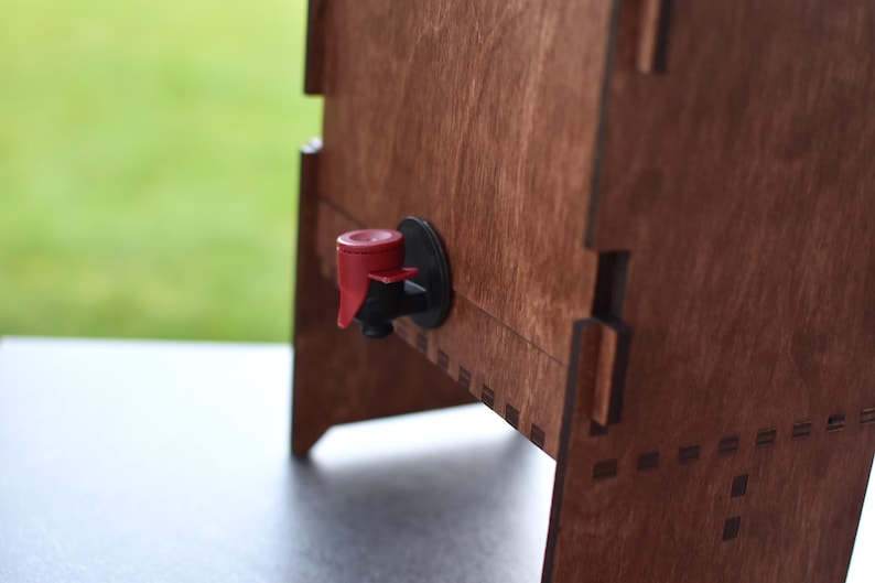 Puede incluir: Un dispensador de madera con un grifo rojo y negro. El dispensador est&aacute; hecho de madera y tiene un acabado natural.