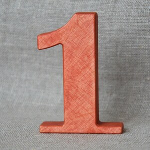 Op de afbeelding: Een houten nummer &eacute;&eacute;n, geverfd in een roodachtig-oranje kleur. Het nummer staat rechtop op een getextureerd oppervlak.
