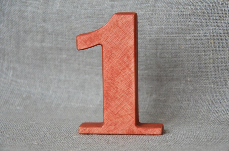 Op de afbeelding: Een houten nummer &eacute;&eacute;n, geverfd in een roodachtig-oranje kleur. Het nummer staat rechtop op een getextureerd oppervlak.