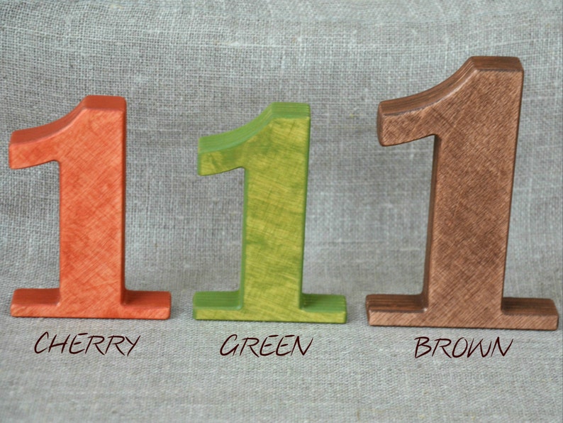 Op de afbeelding: Drie houten nummers &eacute;&eacute;n, geschilderd in verschillende kleuren: kersrood, groen en bruin. De nummers staan op een getextureerd oppervlak.
