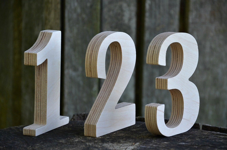 Peut inclure: Trois chiffres en bois, 1, 2 et 3, sont dispos&eacute;s en rang&eacute;e. Les chiffres sont en contreplaqu&eacute; et ont une finition peinte blanche. Les chiffres sont pos&eacute;s sur une surface en bois.