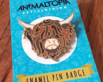 May include: Enamel pin badge featuring a Highland Cow head with brown and white fur. The pin is gold and has a blue background with the text "Animaltopia Art + Clothing" and "myanimaltopia.com".