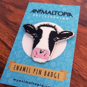 May include: A black and white enamel pin badge featuring a cow's face with a pink nose. The pin is on a blue background with white text that reads "Enamel Pin Badge" and "myanimaltopia.com".
