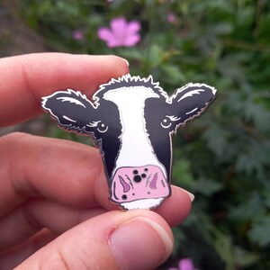 May include: A black and white enamel pin of a cow's face with a pink nose. The pin is held in a hand against a background of green leaves and pink flowers.