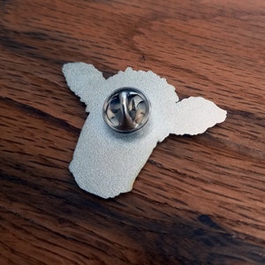 May include: Silver metal pin of a cartoon cow with a white face and black spots. The pin has a silver metal back with a safety pin closure.