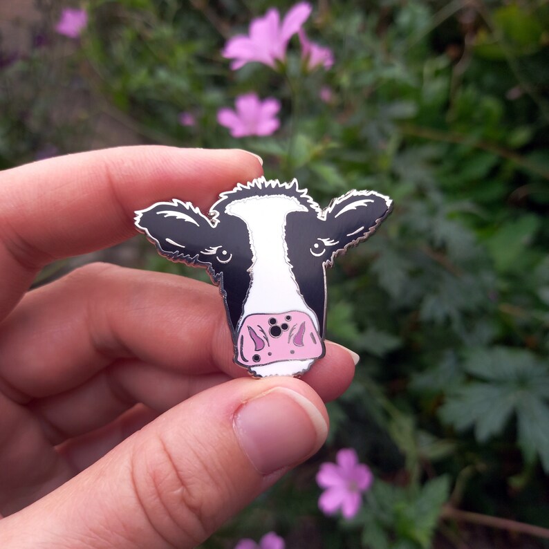 May include: A black and white enamel pin of a cow's face with a pink nose. The pin is held in a hand against a background of green leaves and pink flowers.