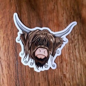 May include: A sticker of a Highland cow with brown fur and a white face. The cow is looking to the right and has a long, shaggy mane.