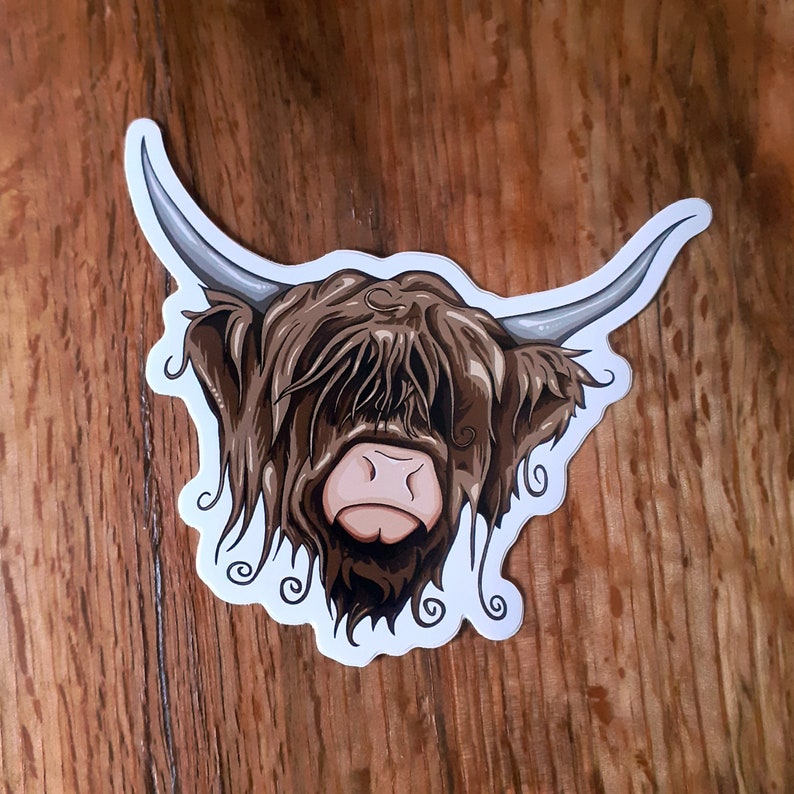 May include: A sticker of a Highland cow with brown fur and a white face. The cow is looking to the right and has a long, shaggy mane.