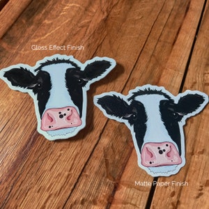 K&ouml;nnte beinhalten: Zwei schwarz-wei&szlig;e Kuh-Aufkleber, einer mit gl&auml;nzendem Finish und der andere mit mattem Papier-Finish. Die Aufkleber befinden sich auf einer Holzoberfl&auml;che.