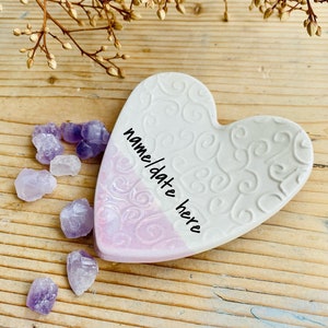 Può includere: Un piatto di ceramica bianco a forma di cuore con un design a spirale rosa e bianco. Il piatto presenta un motivo a spirale decorativo e le parole "name/date here" scritte su di esso. Il piatto è circondato da piccoli cristalli viola.