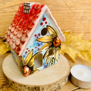 Könnte beinhalten: Ein Kerzenhalter aus Keramik in Form eines Hauses mit einem roten Dach und einem blauen und weißen Blumendesign. Das Haus hat ein Fenster und einen kleinen Schornstein. Der Kerzenhalter steht auf einer Holzoberfläche.