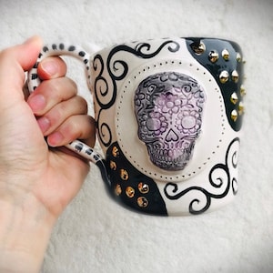 Pode incluir: Caneca de cer&acirc;mica branca com um design em preto e branco que apresenta uma caveira de a&ccedil;&uacute;car com detalhes em roxo e branco. A caneca tem uma al&ccedil;a preta com bolinhas brancas e tachas douradas ao redor da borda.