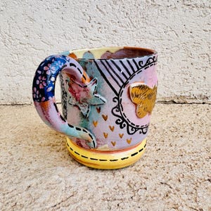 May include: A hand-painted ceramic mug with a colorful, abstract design. The mug features a blue handle with floral patterns, a yellow base, and a body adorned with stars, hearts, and a gold-colored emblem. The mug is approximately 4 inches tall.