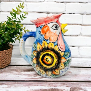 Op de afbeelding: Een keramische kan in de vorm van een haan met een zonnebloemdessin. De kan is geschilderd in blauw, geel, oranje, rood en wit.