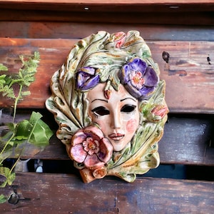 Peut inclure: Un masque en céramique représentant un visage de femme, décoré de fleurs et de feuilles. Le masque est peint dans des tons de vert, rose, violet et or.