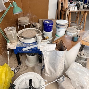 May include: A pottery studio workspace with a pottery wheel, a variety of buckets, bags of clay, and a drill. There are also several ceramic pieces on a shelf in the background.