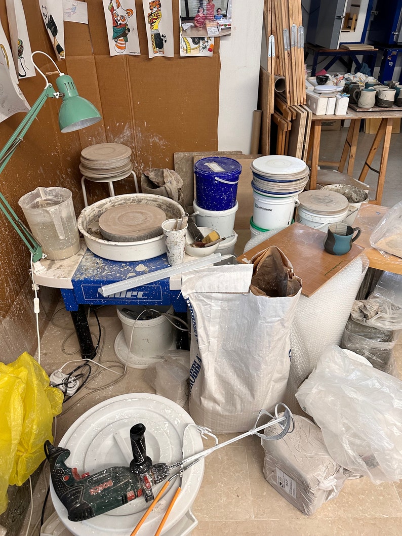 May include: A pottery studio workspace with a pottery wheel, a variety of buckets, bags of clay, and a drill. There are also several ceramic pieces on a shelf in the background.