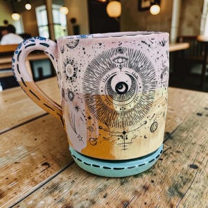 Pode incluir: Uma caneca de cerâmica com um design celestial apresentando um motivo de olho central e lua crescente. A caneca tem uma seção superior rosa claro, uma seção inferior amarelo mostarda e uma base azul claro. A alça é decorada com padrões azuis.