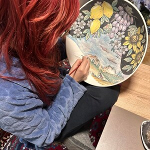May include: A woman with red hair paints a ceramic plate with a colorful landscape scene. The plate features lemons, grapes, sunflowers, and a mountain range.