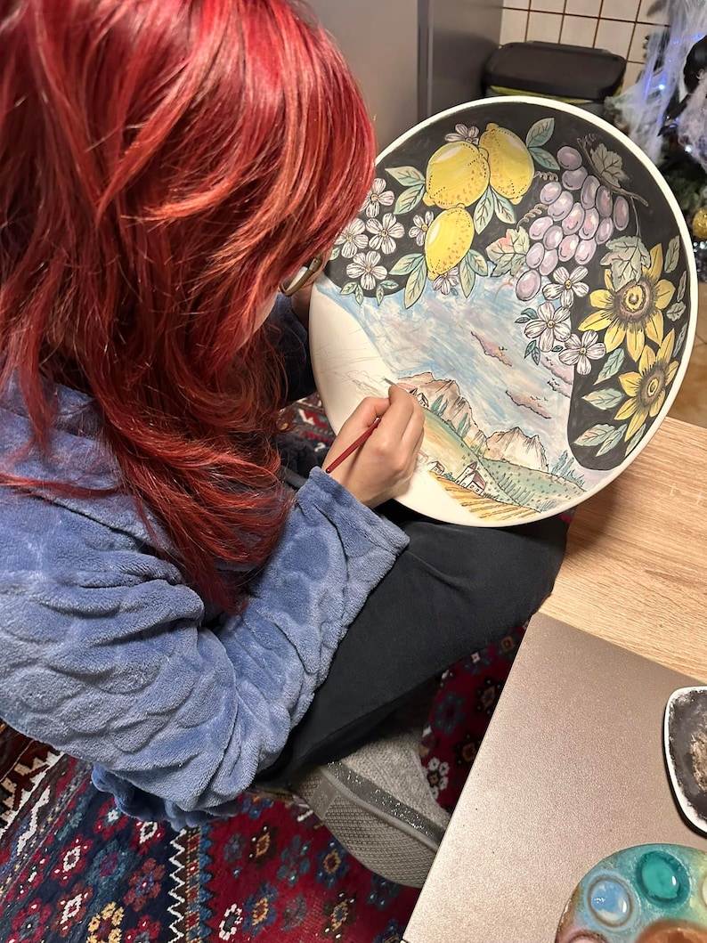 May include: A woman with red hair paints a ceramic plate with a colorful landscape scene. The plate features lemons, grapes, sunflowers, and a mountain range.