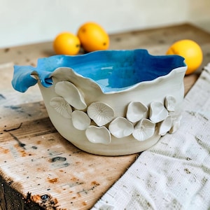Op de afbeelding: Een handgemaakte keramische kom met een gladde, gebroken witte buitenkant en een felblauwe binnenkant. De buitenkant is versierd met verschillende kleine, verhoogde, witte bloembladachtige vormen. De kom heeft een onregelmatige, golvende rand.