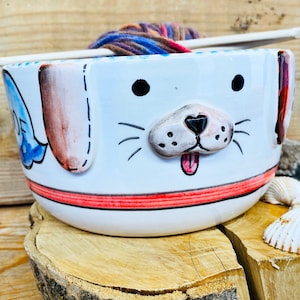 May include: White ceramic yarn bowl shaped like a dog's face with a red stripe around the rim. The bowl has a brown nose, black eyes, and a pink tongue. A ball of yarn is resting on the rim of the bowl.