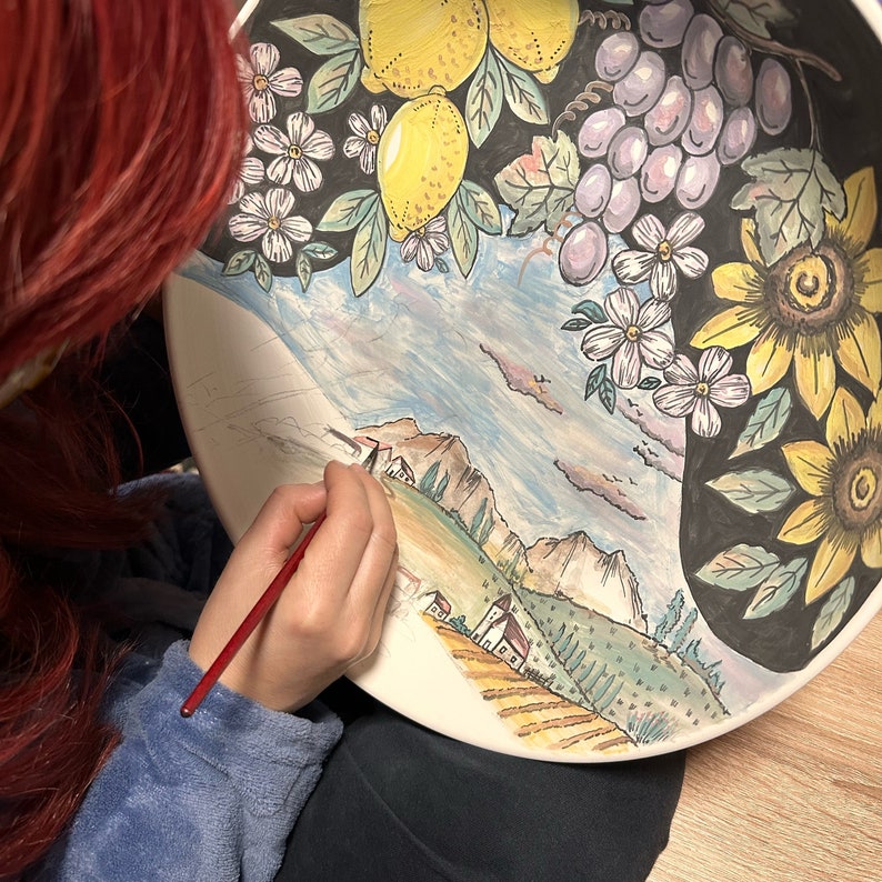 Puede incluir: Un plato grande de cer&aacute;mica con un paisaje pintado a mano de monta&ntilde;as, campos y casas peque&ntilde;as. El borde est&aacute; decorado con limones amarillos, uvas moradas, flores blancas y girasoles amarillos sobre un fondo de color negro.