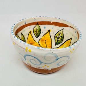 May include: A white ceramic bowl with a hand-painted design of yellow leaves and blue swirls. The bowl has a brown rim and yellow dots.