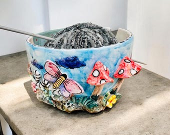 May include: A ceramic yarn bowl featuring a light blue sky background with hand-painted details including a pink butterfly, two red mushrooms with white spots, and small yellow flowers. A ball of grey yarn sits inside with a metal knitting needle.