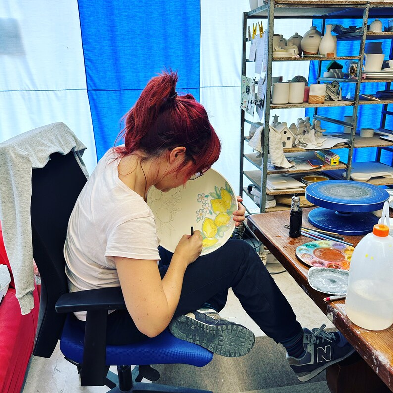 May include: A person painting a white ceramic plate with yellow lemons and white flowers. The plate is on a table with a paint palette and brushes.