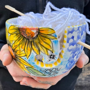May include: White yarn is wound around a ceramic yarn bowl with a sunflower and bee design. The bowl has a blue and white pattern on the side and a yellow and white honeycomb pattern on the bottom.