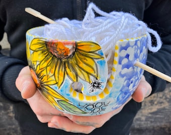 May include: White yarn is wound around a ceramic yarn bowl with a sunflower and bee design. The bowl has a blue and white pattern on the side and a yellow and white honeycomb pattern on the bottom.