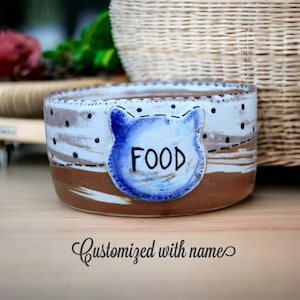 May include: A ceramic pet food bowl with a brown and white speckled glaze. The bowl has a blue cat-shaped design with the word "FOOD" written on it. The bowl is sitting on a wooden surface.