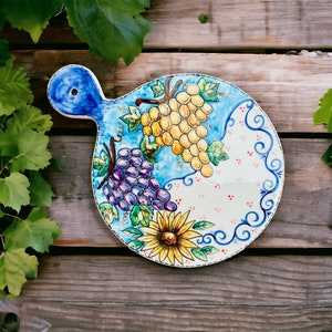 Pode incluir: Um prato de servir redondo de cerâmica com um design azul, branco e amarelo. O prato apresenta uvas, um girassol e uma alça azul.