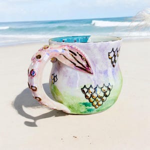 May include: A ceramic mug with a mermaid tail handle. The mug is painted with a purple, green, and blue ombre design and features gold glitter scales. The handle is a pink and white mermaid tail with gold accents.