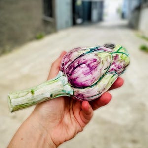 Peut inclure: Une main tient une pipe en céramique en forme d'artichaut. La pipe est peinte avec des détails de type aquarelle en violet, vert et blanc. La pipe a un petit trou sur le dessus pour fumer.