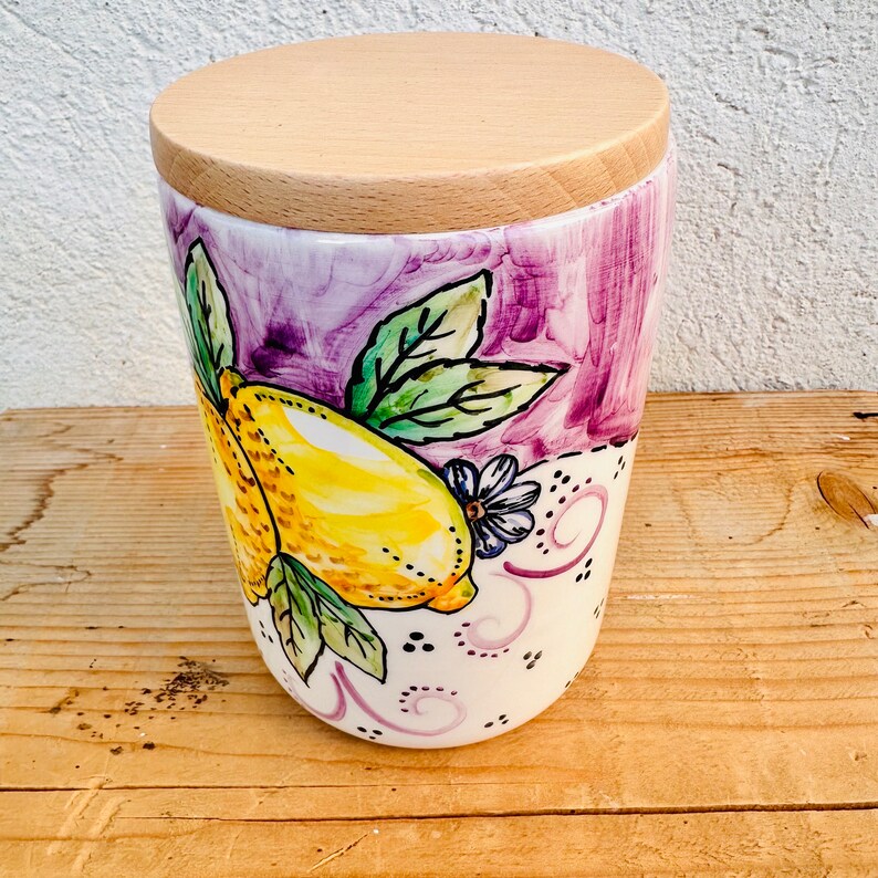 Puede incluir: Un recipiente de cer&aacute;mica con tapa de madera clara que presenta un dise&ntilde;o pintado a mano de limones amarillos con hojas verdes y trazos morados. La base blanca est&aacute; decorada con peque&ntilde;os puntos negros y espirales de color morado.