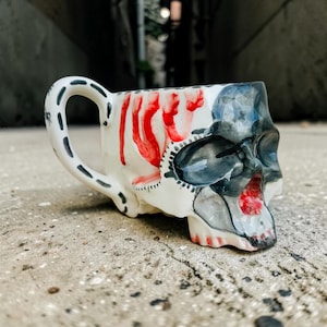 Peut inclure: Une tasse en céramique en forme de crâne humain avec une anse. La tasse présente une base blanche avec des ombres gris foncé autour des orbites et de la cavité nasale, des gouttes de peinture rouge vif sur le côté et un intérieur rouge.