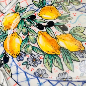 May include: A white ceramic plate with a blue and white checkered border. The plate is decorated with a hand-painted design of lemons, olives, and flowers. The lemons are yellow with green leaves, the olives are green, and the flowers are purple.