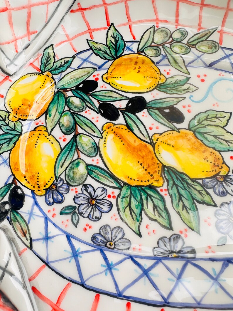 May include: A white ceramic plate with a blue and white checkered border. The plate is decorated with a hand-painted design of lemons, olives, and flowers. The lemons are yellow with green leaves, the olives are green, and the flowers are purple.