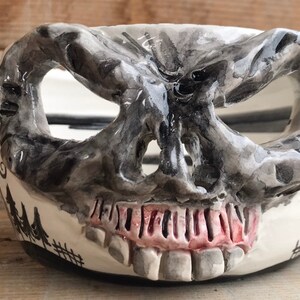 May include: A ceramic bowl shaped like a skull with a black and white painted design. The bowl has a wide, open mouth and a set of teeth. The bowl is painted with a forest scene on the side.