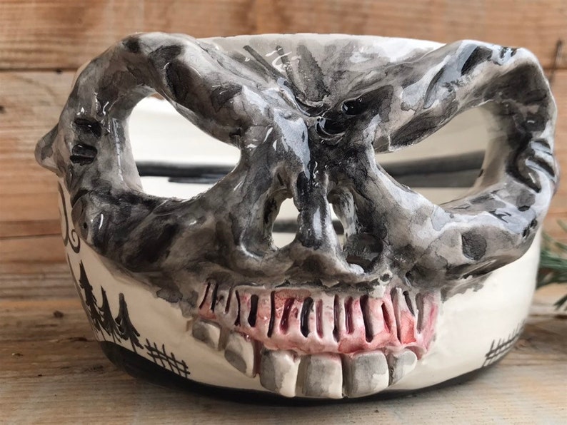 May include: A ceramic bowl shaped like a skull with a black and white painted design. The bowl has a wide, open mouth and a set of teeth. The bowl is painted with a forest scene on the side.