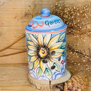 May include: A ceramic garlic keeper featuring a sunflower design. The lid is blue and white with the word "Garlic" written on it. The keeper is decorated with a yellow sunflower with a brown centre and green leaves. The background is blue with white swirls.