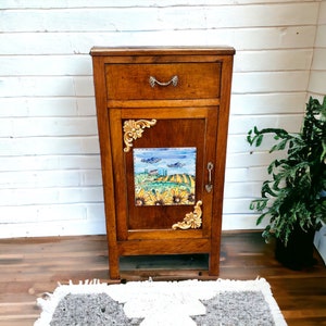 May include: A wooden cabinet with a single drawer and a door. The door features a decorative tile with a painted scene of a countryside landscape with sunflowers. The cabinet is brown with gold accents.