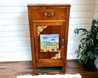 May include: A wooden cabinet with a single drawer and a door. The door features a decorative tile with a painted scene of a countryside landscape with sunflowers. The cabinet is brown with gold accents.
