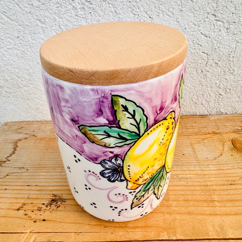 Puede incluir: Un recipiente de cer&aacute;mica con tapa de madera clara que presenta un dise&ntilde;o pintado a mano de limones amarillos brillantes y hojas verdes sobre un fondo de color violeta. La parte inferior del recipiente blanco tiene puntos negros.