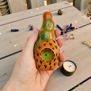 Könnte beinhalten: Eine handbemalte Keramikpfeife mit einem orange-grünen Design, verziert mit schwarzen Herzformen. Die Pfeife hat einen grünen Kopf und wird von einem kleinen, grünen Keramikbehälter mit weißem Innenraum begleitet.