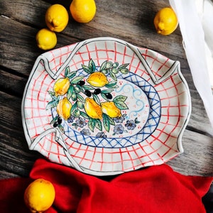 May include: A white ceramic serving platter with a red and blue checkered pattern. The platter features a hand-painted design of lemons and olives on a branch with green leaves.