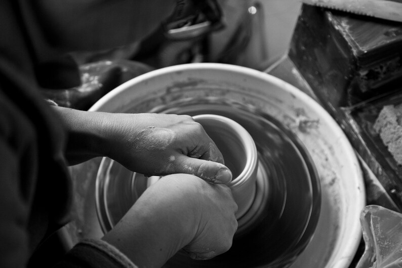 May include: A person's hands shaping a clay pot on a pottery wheel. The wheel is spinning and the clay is being molded into a round shape.